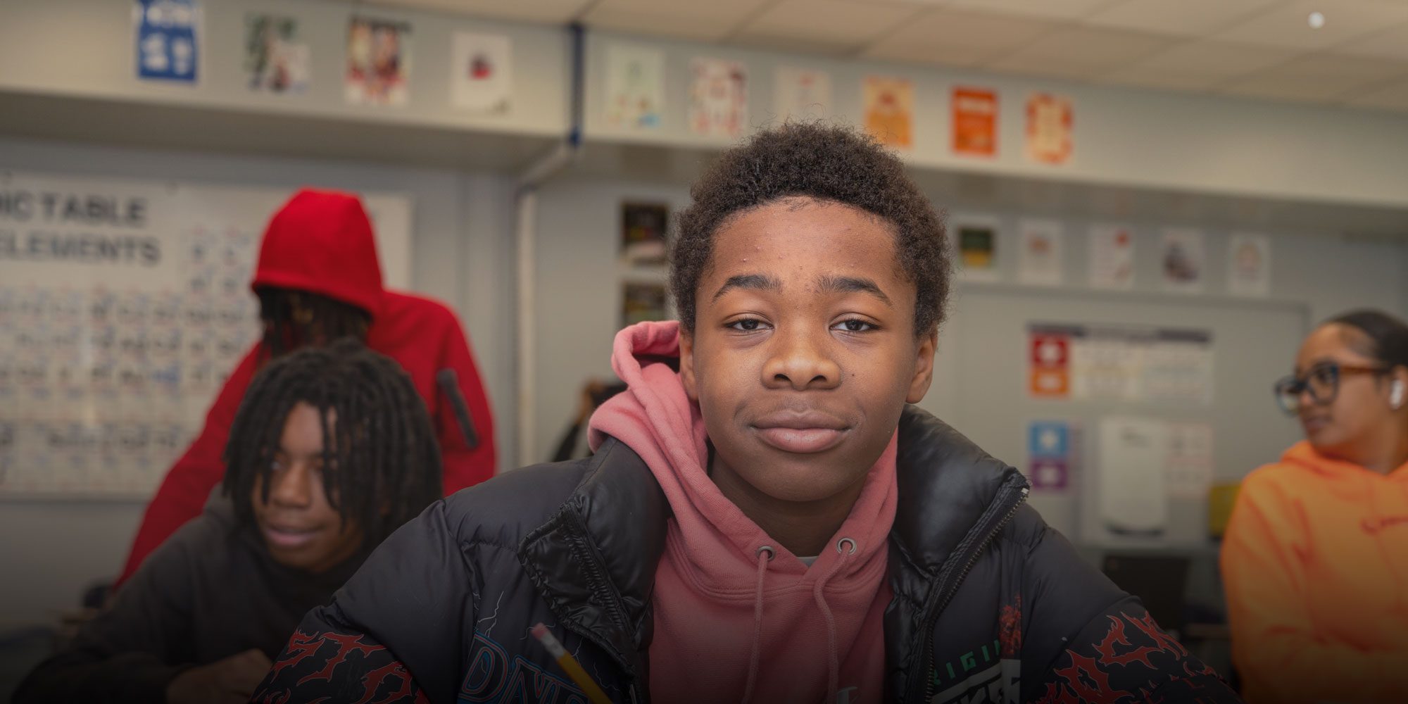 Student in a pink hoodie and black jacket in a classroom, with classmates in the background.