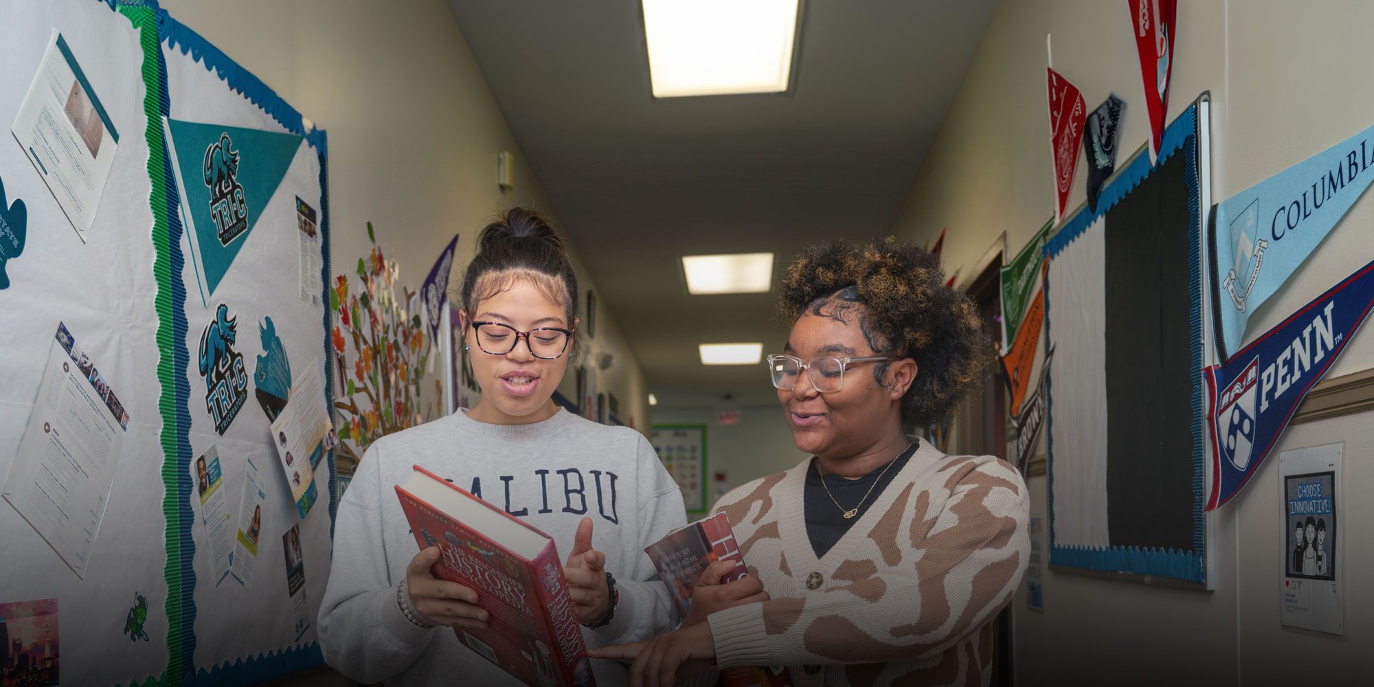 Two people look through books in a school hallway lined with college pennants.